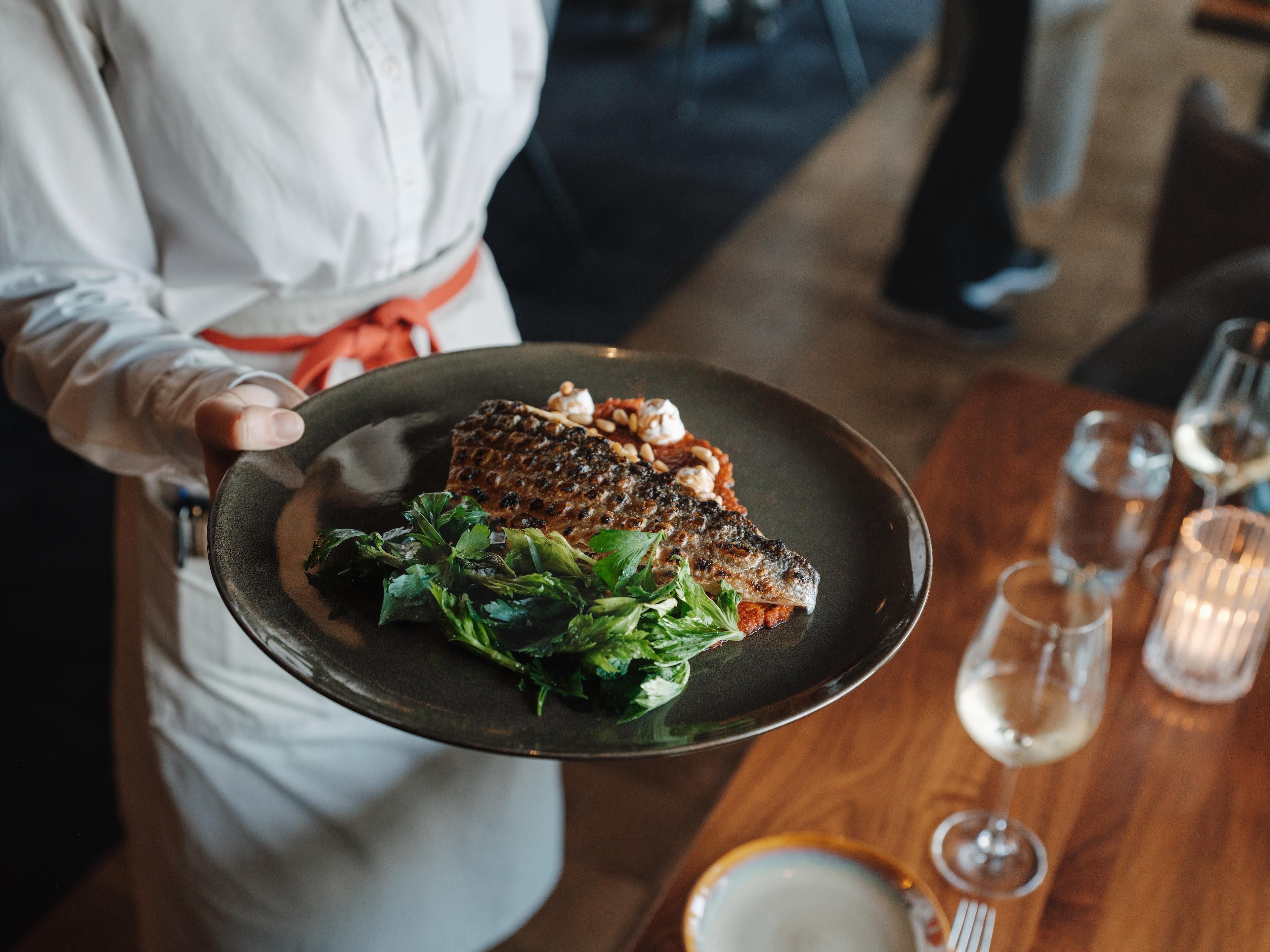Une personne tenant une assiette avec du poisson grillé et des légumes verts, un verre de vin blanc et une bougie sur une table en bois.