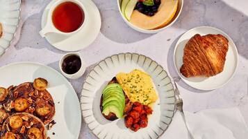 Un desayuno abundante con tortitas, huevos, tostadas y fruta en platos.