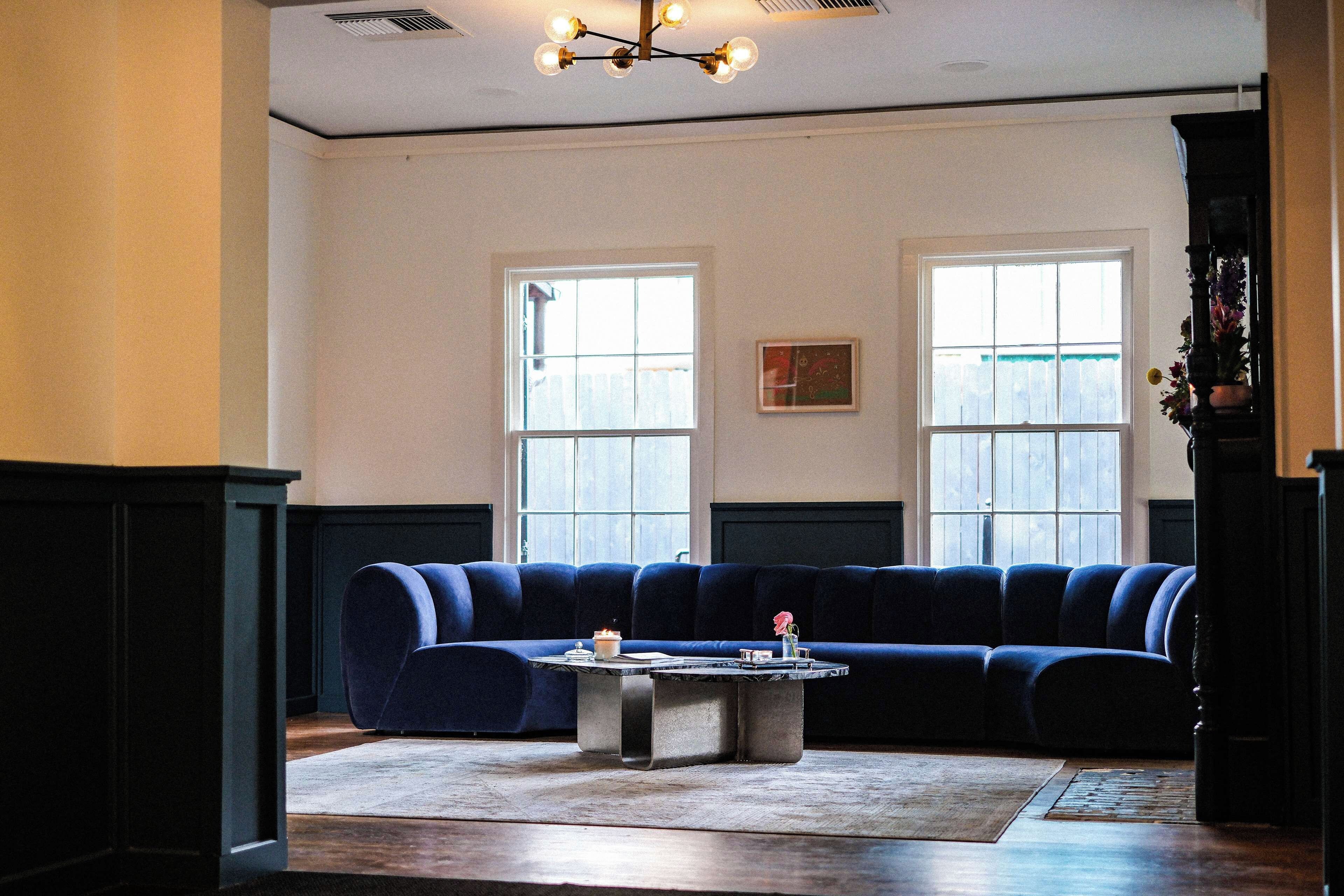 A living room with a dark blue sectional sofa, a round coffee table, and a chandelier. There are two windows with white frames and a painting on the wall.