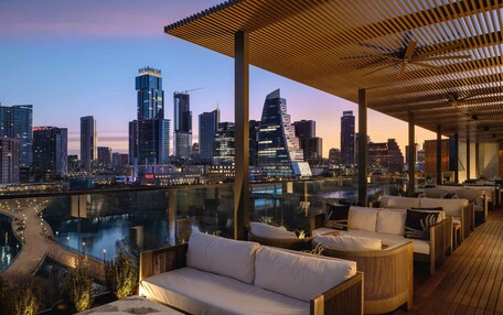 A rooftop lounge with a city view, modern furniture, and a glass railing overlooking a river.