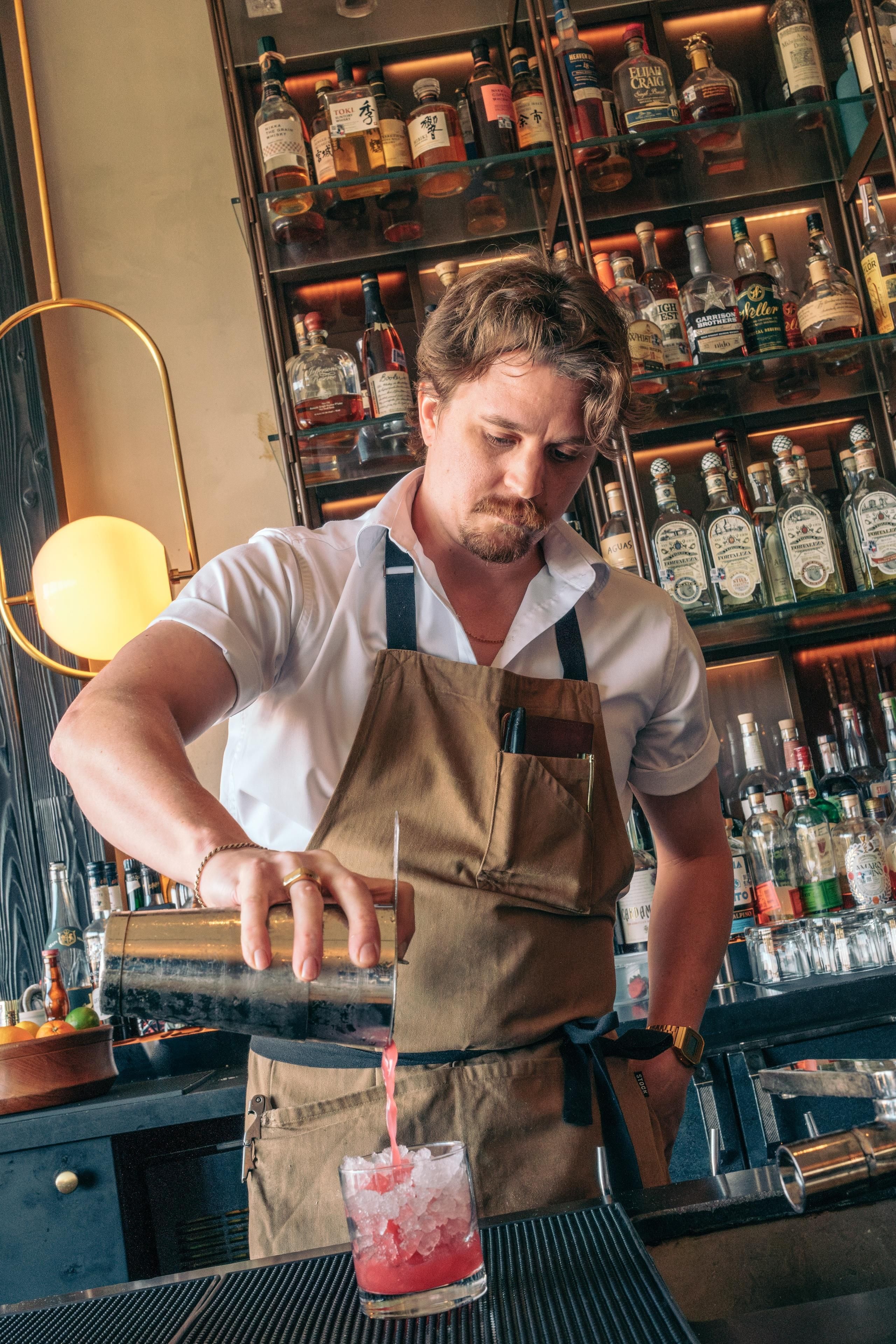 Un bartender preparando una bebida en la barra, con estantes llenos de botellas de licor al fondo.