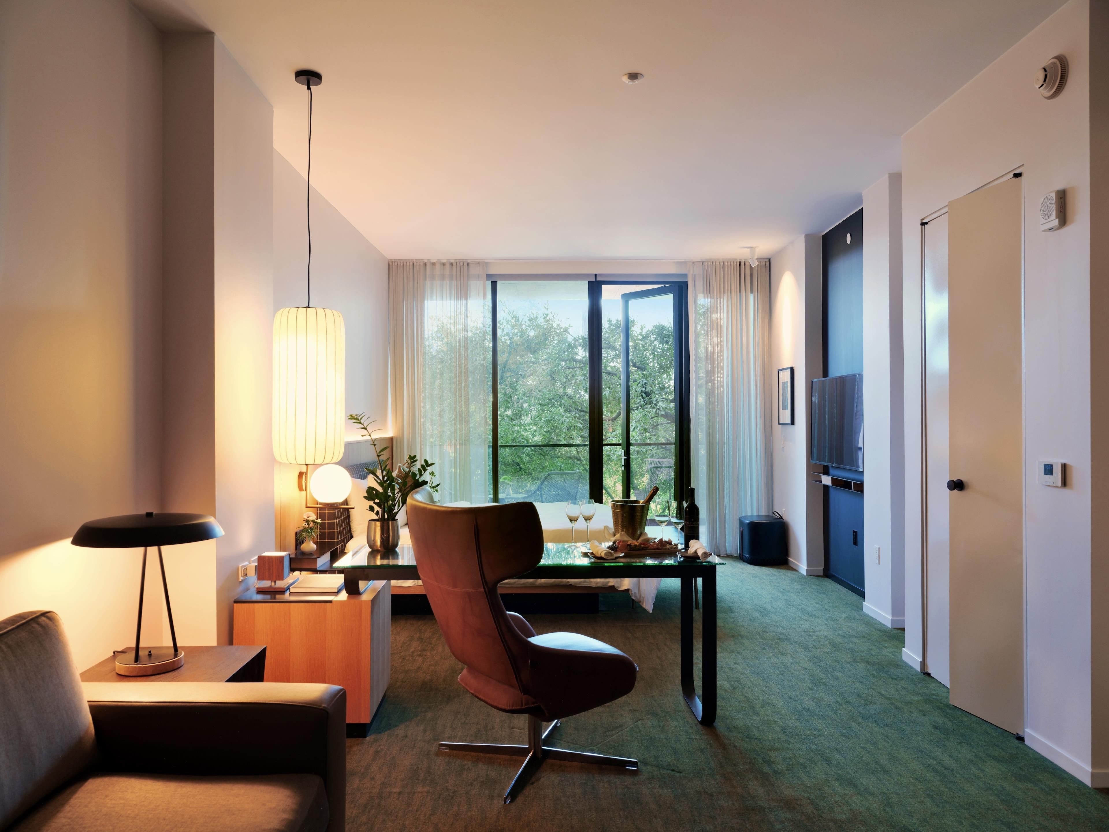 A modern hotel room with a sofa, a dining table with a chair, a TV, and a large window offering a view of trees.