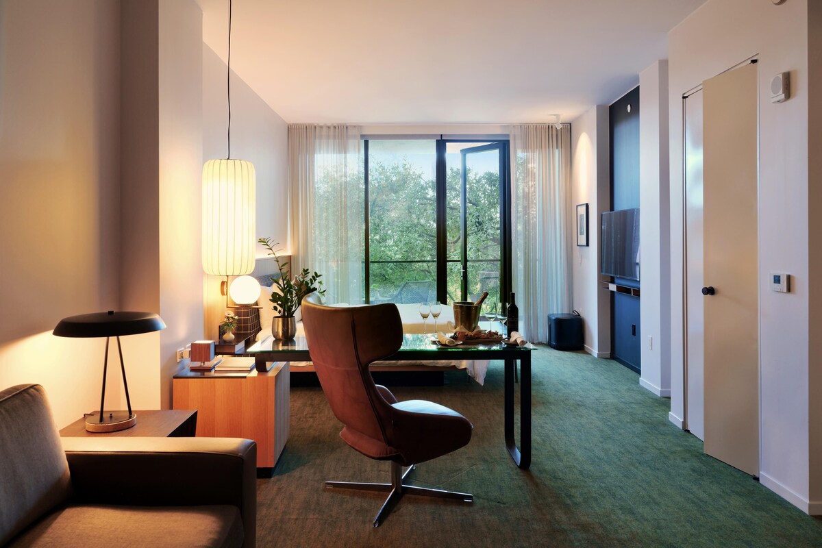 A modern hotel room with a sofa, a dining table with a chair, a TV, and a large window offering a view of trees.