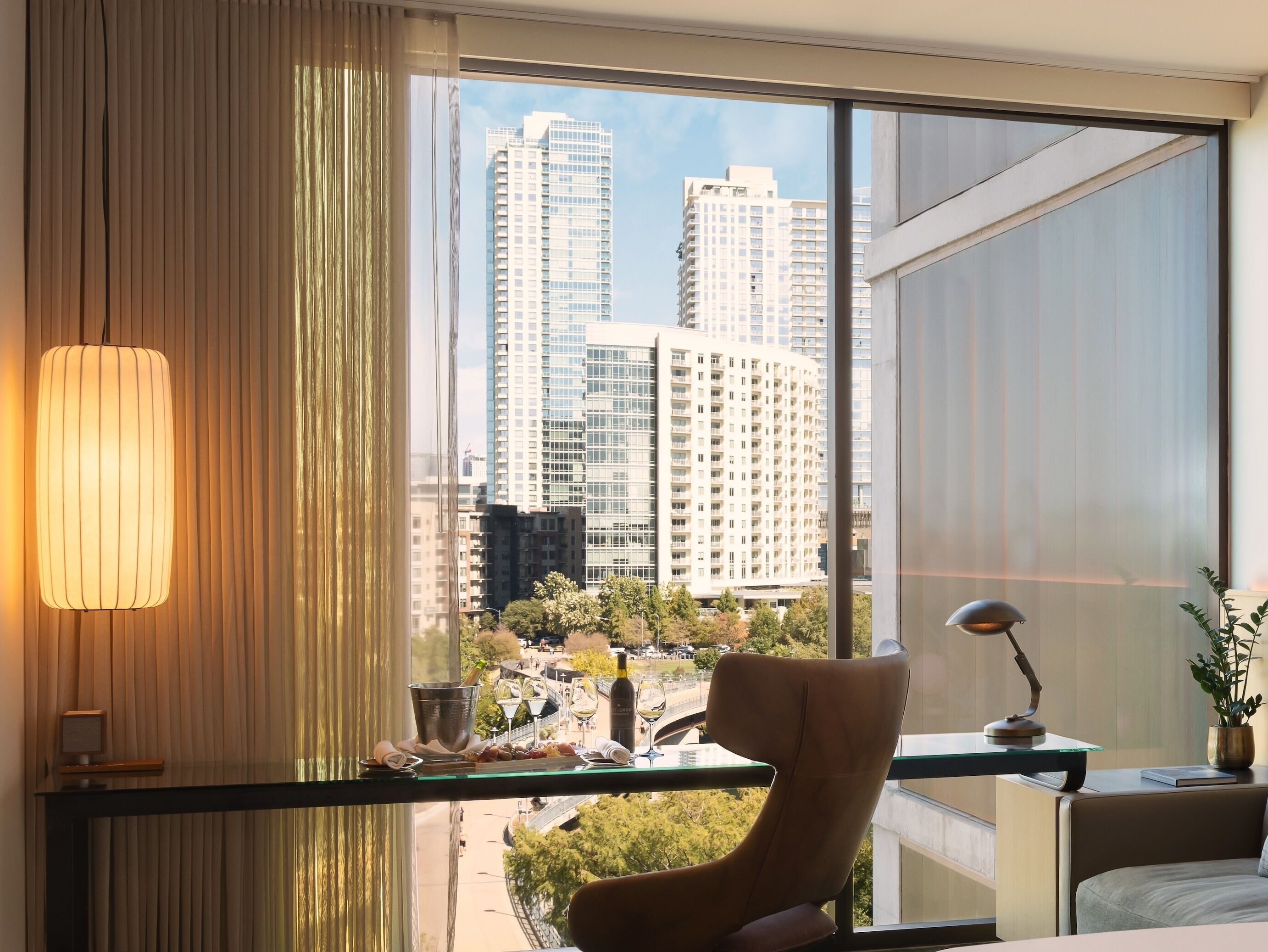 A modern hotel room with a large window offering a city view, a comfortable chair, a small table with a lamp, and a decorative plant.