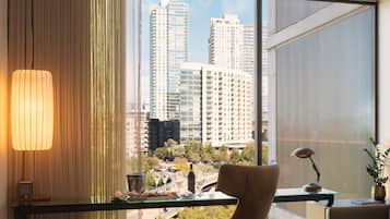 A modern hotel room with a large window offering a city view, a comfortable chair, a small table with a lamp, and a decorative plant.