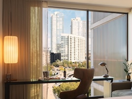 A modern hotel room with a large window offering a city view, a comfortable chair, a small table with a lamp, and a decorative plant.