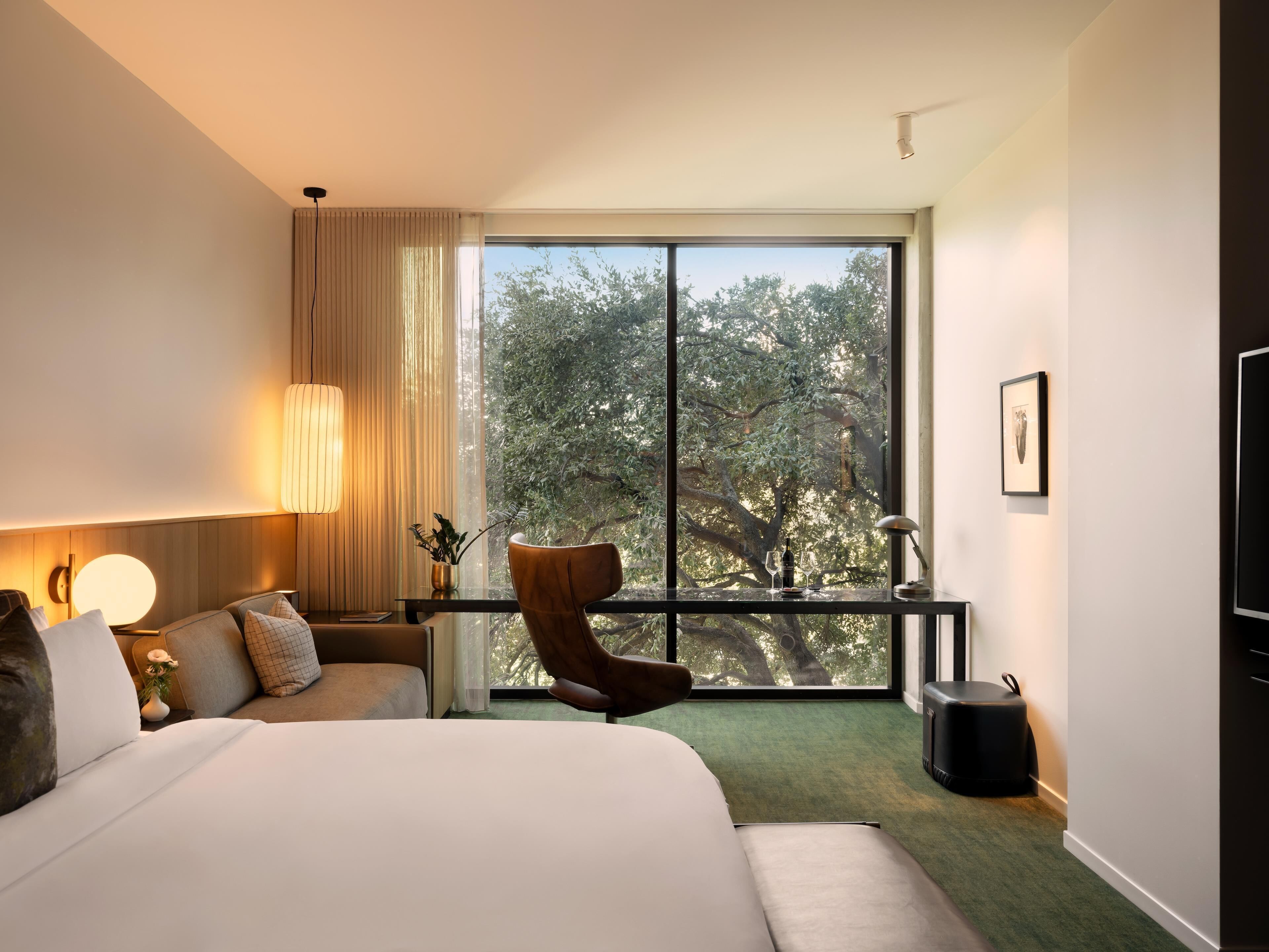 A hotel room with a large bed, a chair, a desk, a lamp, a picture on the wall, and a view of trees through the window.
