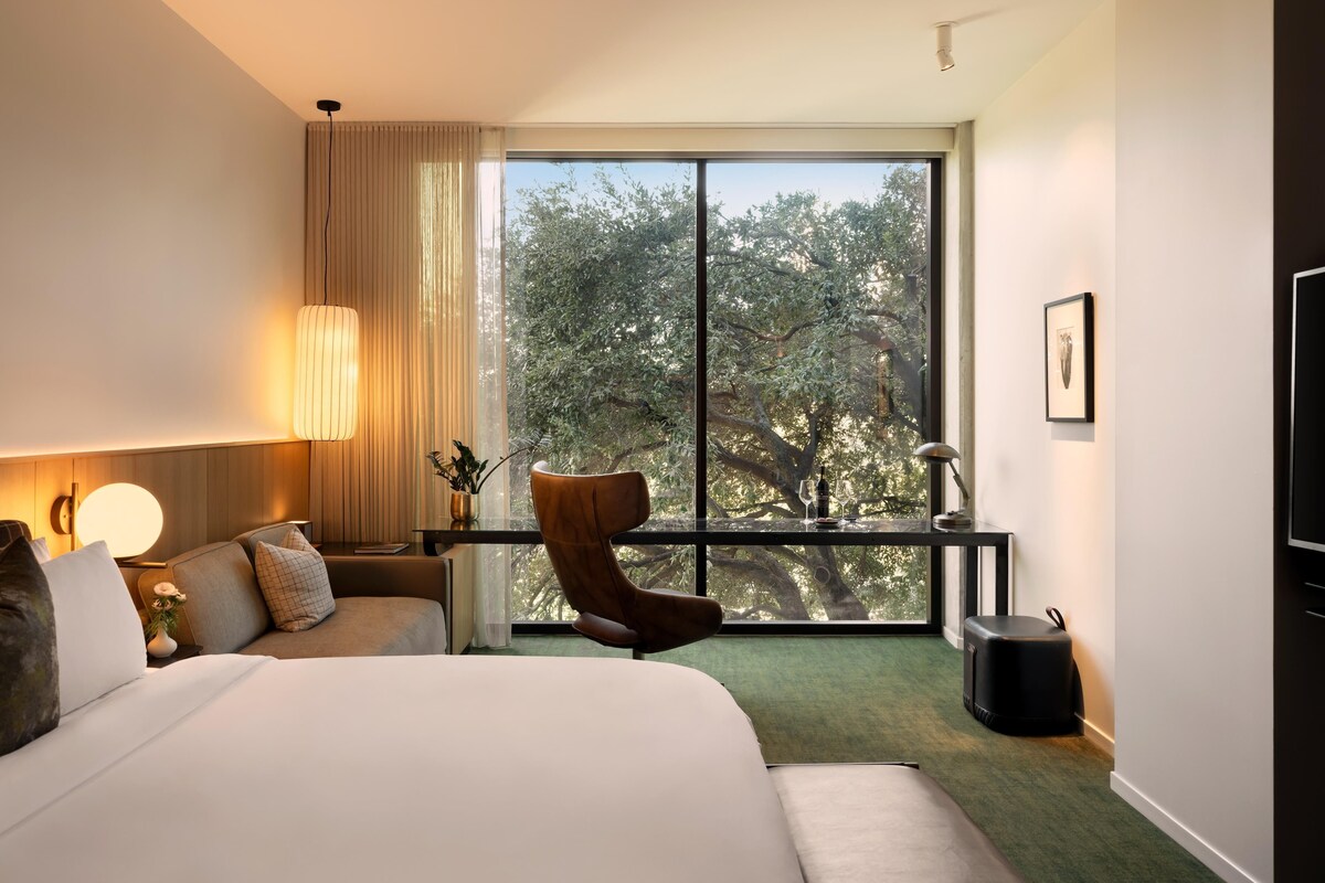 A hotel room with a large bed, a chair, a desk, a lamp, a picture on the wall, and a view of trees through the window.