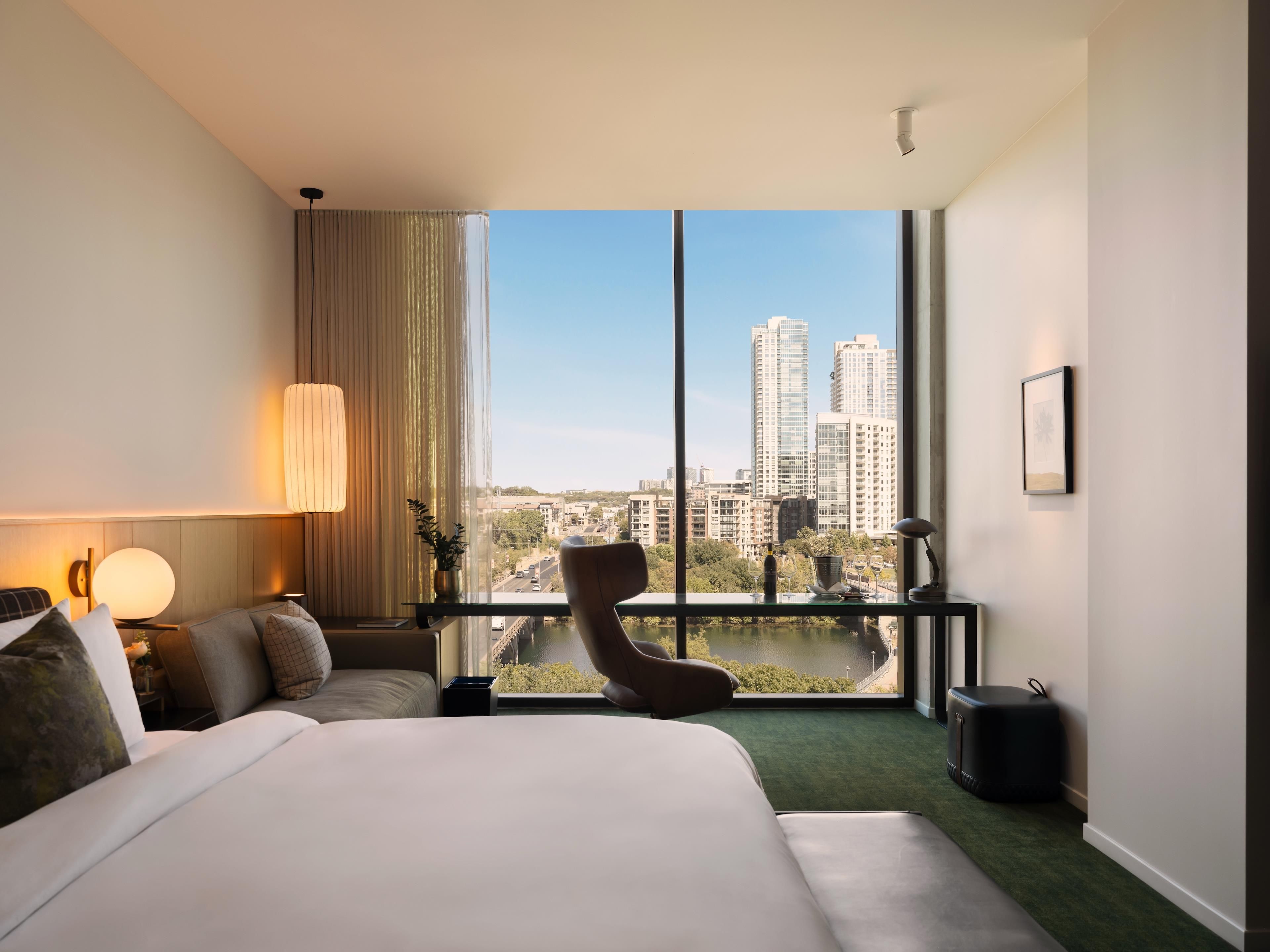 Habitación de hotel con una cama grande, una silla, un escritorio con lámpara y vistas a la ciudad a través de ventanas amplias.