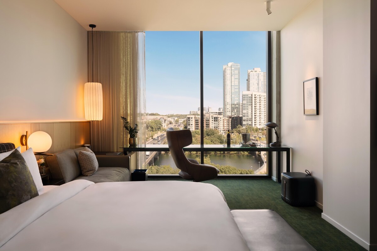 A hotel room with a large bed, a chair, a desk with a lamp, and a view of the cityscape through large windows.