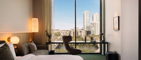 Habitación de hotel con una cama grande, una silla, un escritorio con lámpara y vistas a la ciudad a través de ventanas amplias.