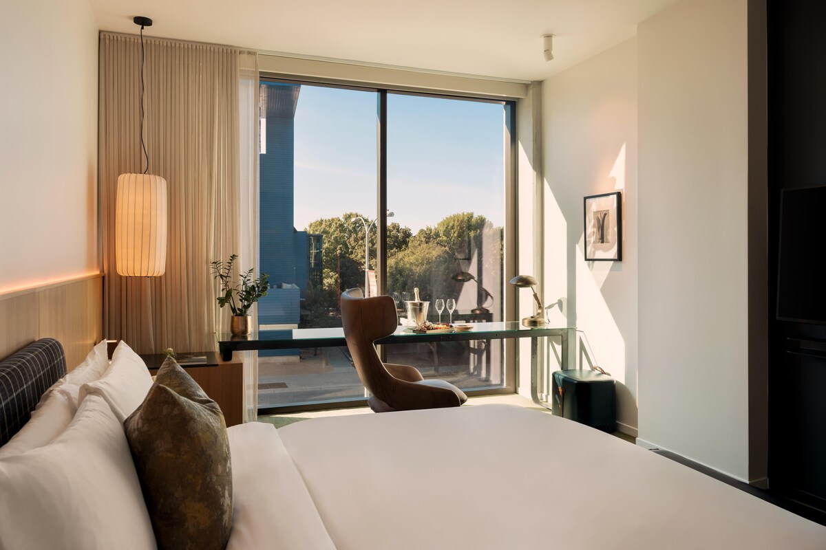 A modern hotel room with a large bed, a sitting area with a chair and table, a large window with a view of a building and trees, and a flat-screen TV on the wall.