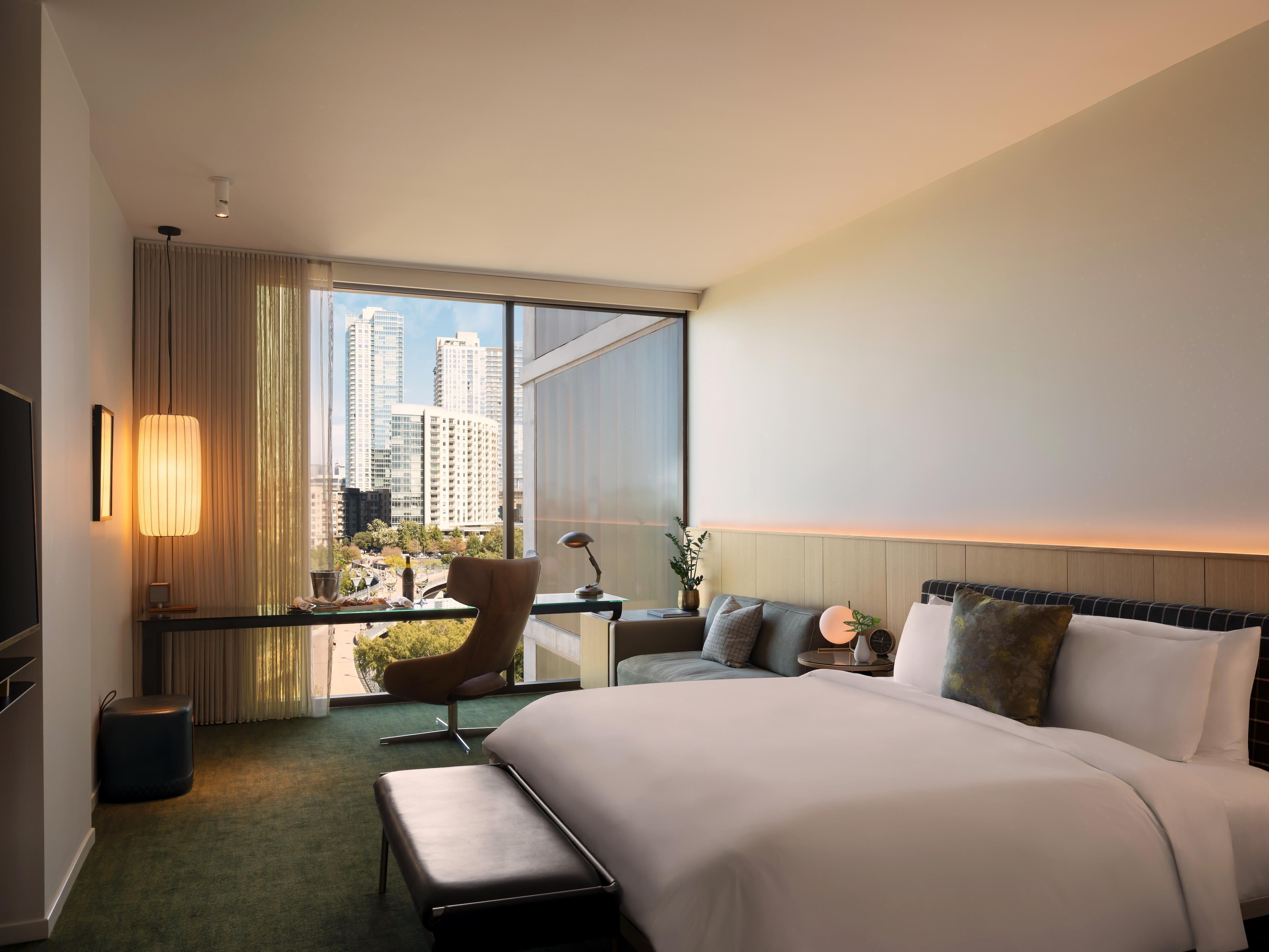 A hotel room with a bed, a desk, a chair, a bench, a TV, and a large window with a city view.