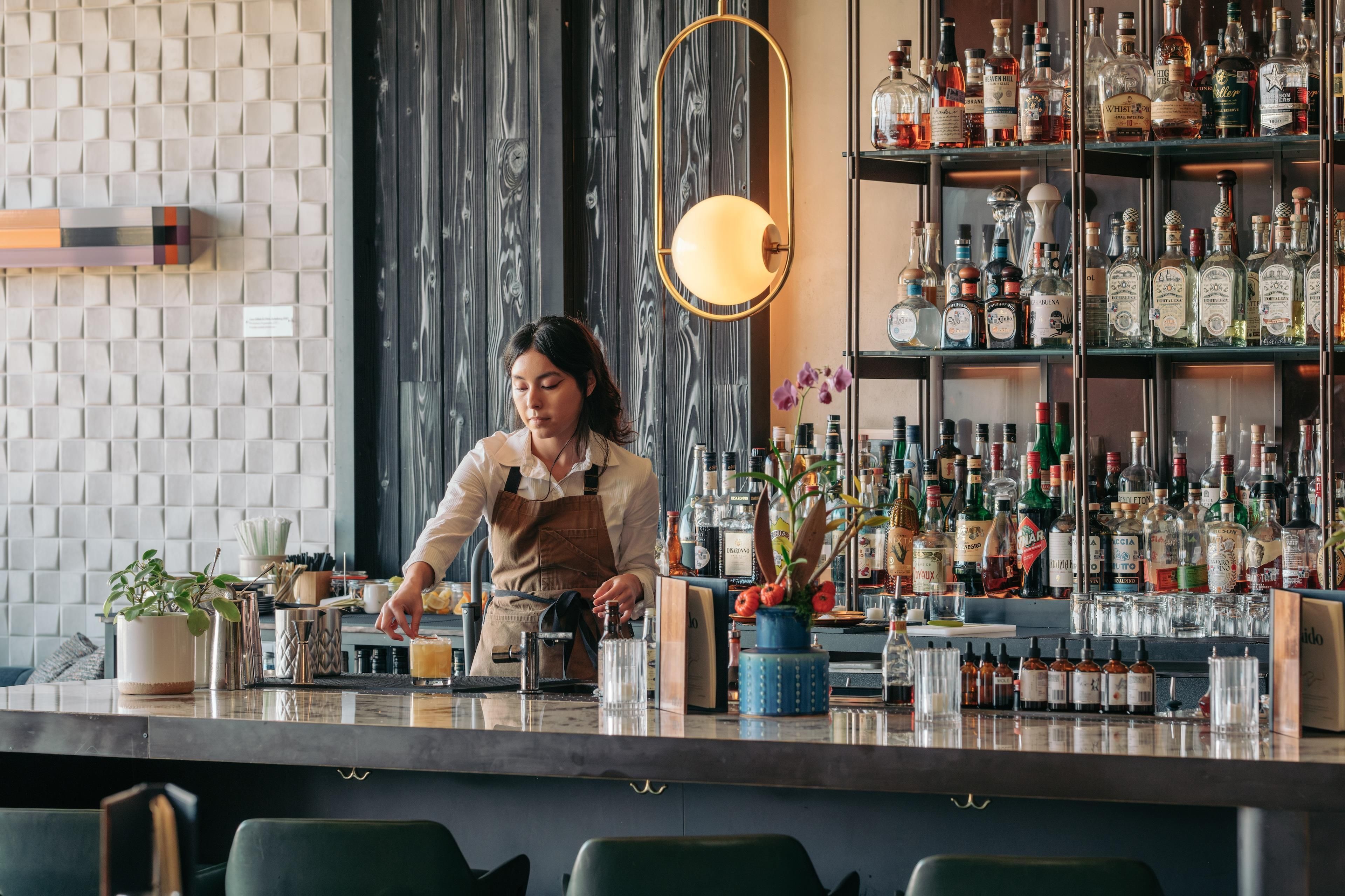 Zona de bar con un barman preparando bebidas, estanterías llenas de diferentes botellas de licor y un diseño de interiores moderno.