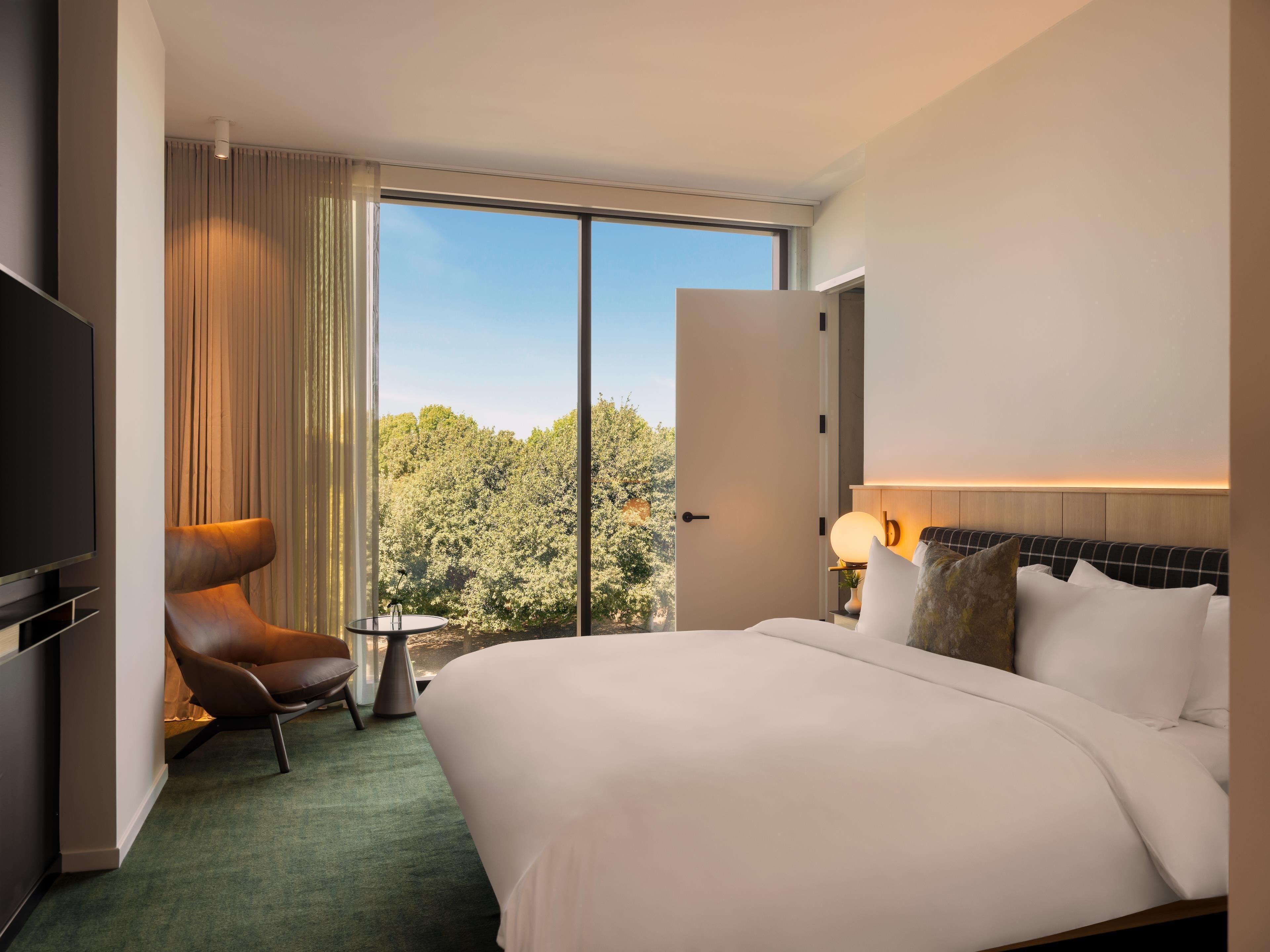A hotel room with a bed, a chair, a small table, a TV, and a view of trees through a large window.