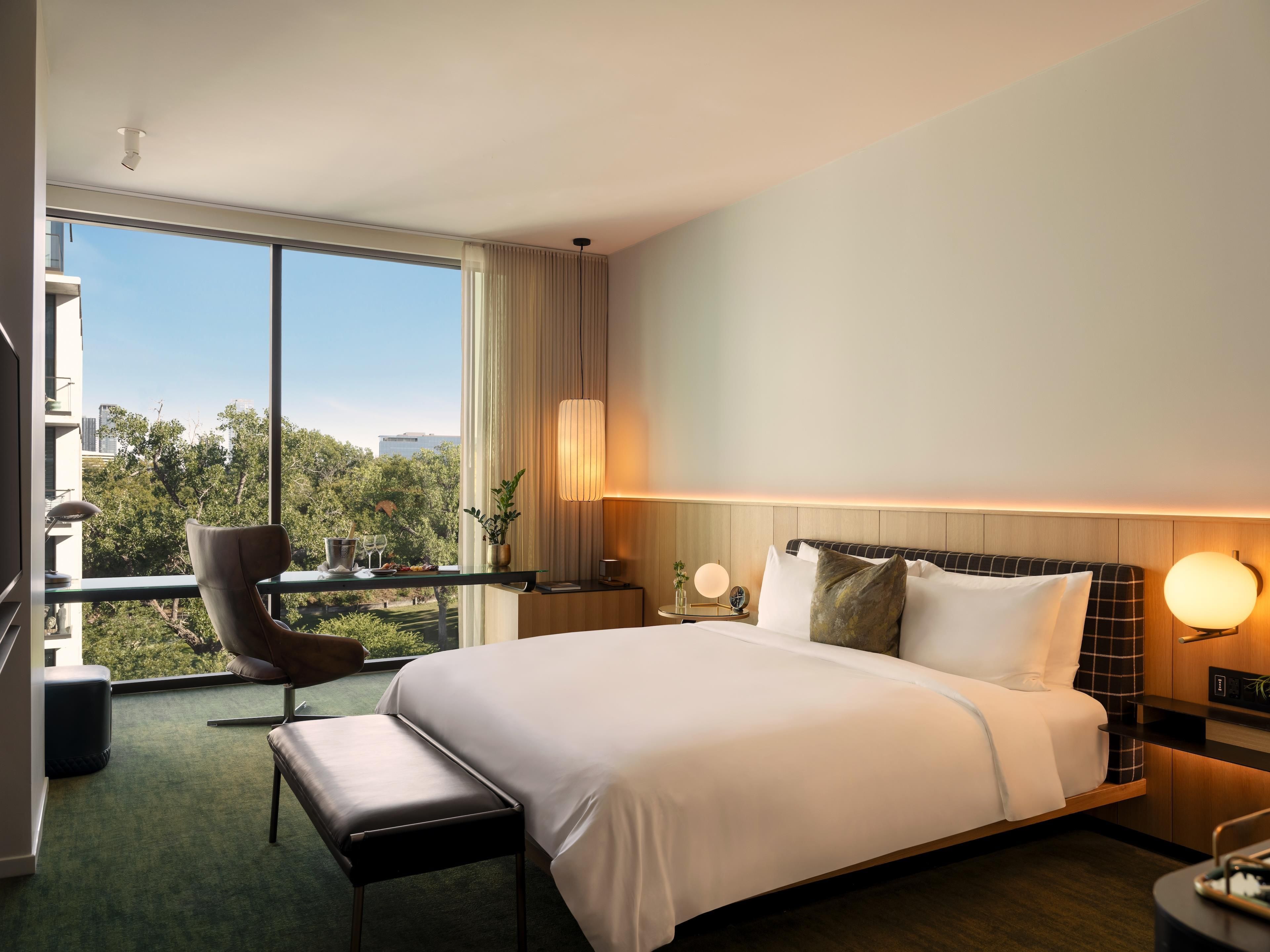 A hotel room with a large bed, a desk with a chair, a view of the outdoors, and a window with curtains.