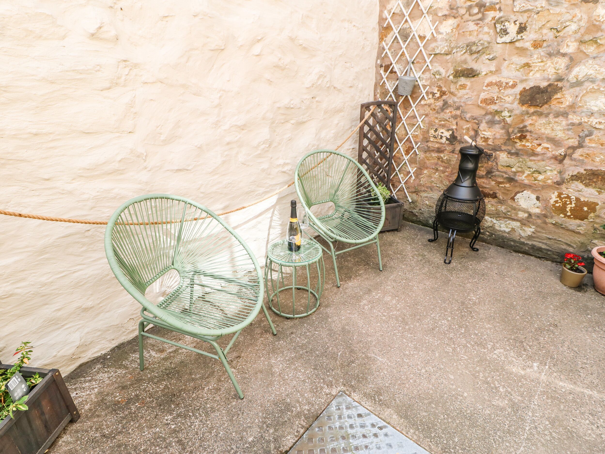 Een terras met twee groene draadstoelen, een tafeltje met een fles en een zwarte metalen heater.