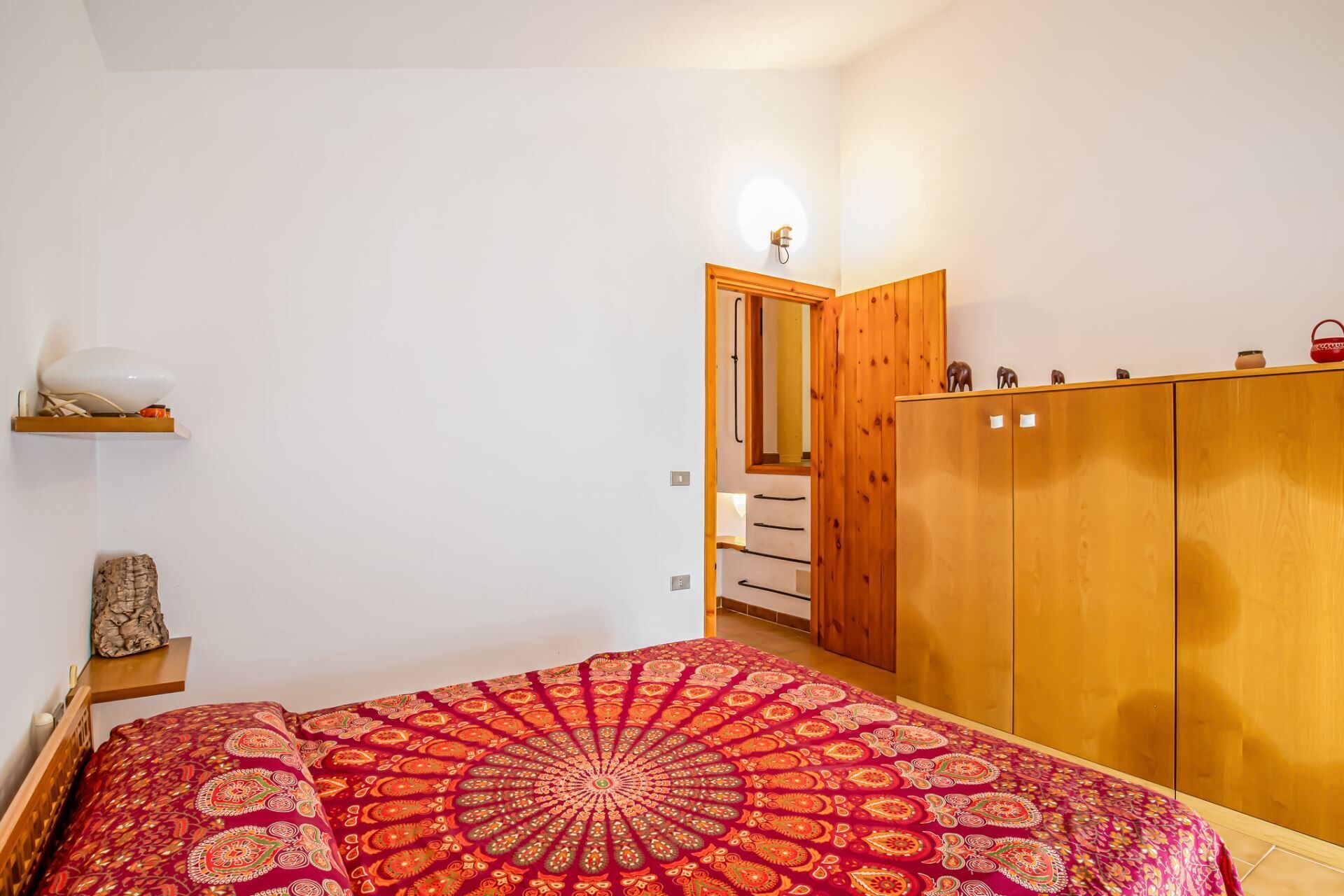 A bedroom with a bed, a wardrobe, a shelf with a lamp, and a door leading to another room.
