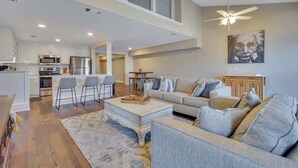 Un vaste espace de vie comprenant un canapé, une table basse et un coin bar. On peut également apercevoir une cuisine équipée d’appareils modernes et un coin repas avec une table et des chaises.