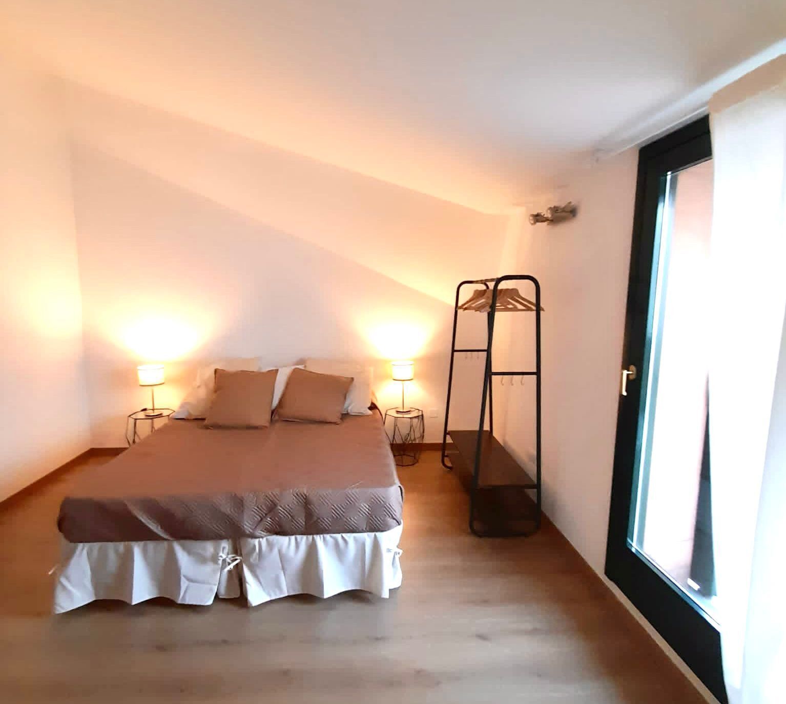 A bedroom with a bed, bedside lamps, a hanging rack, and a large window.