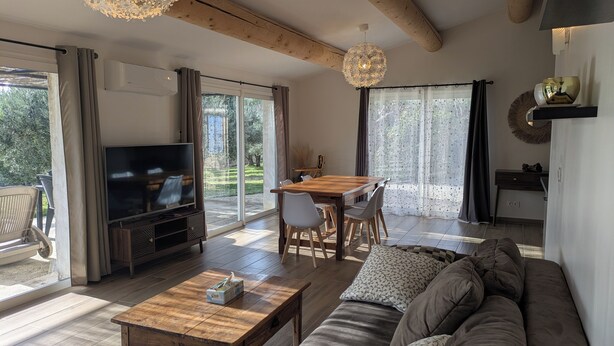 A spacious living room with a wooden coffee table, a sofa, and a TV. There is a dining area with a table and chairs, and large windows offering a view of the outdoors.