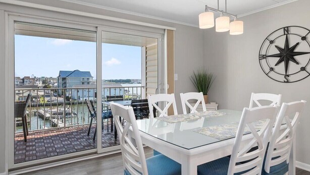 A dining area with a glass table and white chairs, a compass wall decoration, and a view of a marina through large sliding doors. - Thunder Island Condo - Beautiful 2 BR 2 BA Next to Convention Center (Ocean City)