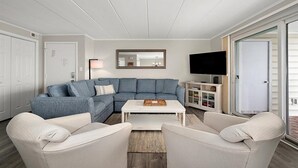A living room with a sectional sofa, a white coffee table, and two armchairs. There is a flat-screen TV mounted on the wall, a mirror above the coffee table, and a cabinet with shelves. - Thunder Island Condo - Beautiful 2 BR 2 BA Next to Convention Center (Ocean City)