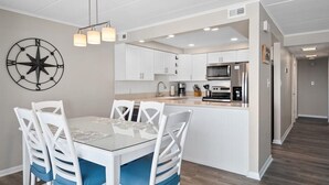 A modern kitchen with white cabinets, a central island, and a dining area with a glass table and white chairs. - Thunder Island Condo - Beautiful 2 BR 2 BA Next to Convention Center (Ocean City)