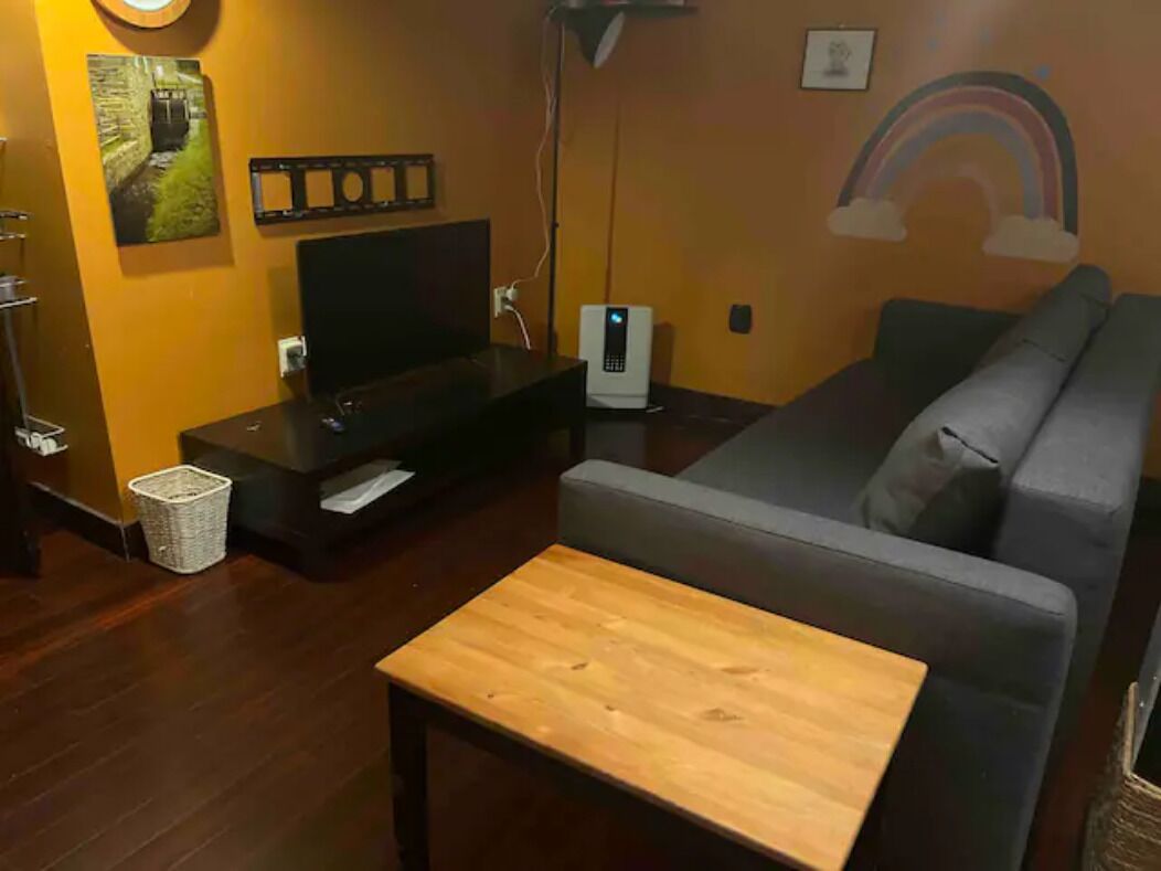 Un soggiorno con un tavolino in legno, un divano grigio, una TV su un mobile, una lampada e una parete con un murale a tema arcobaleno.
