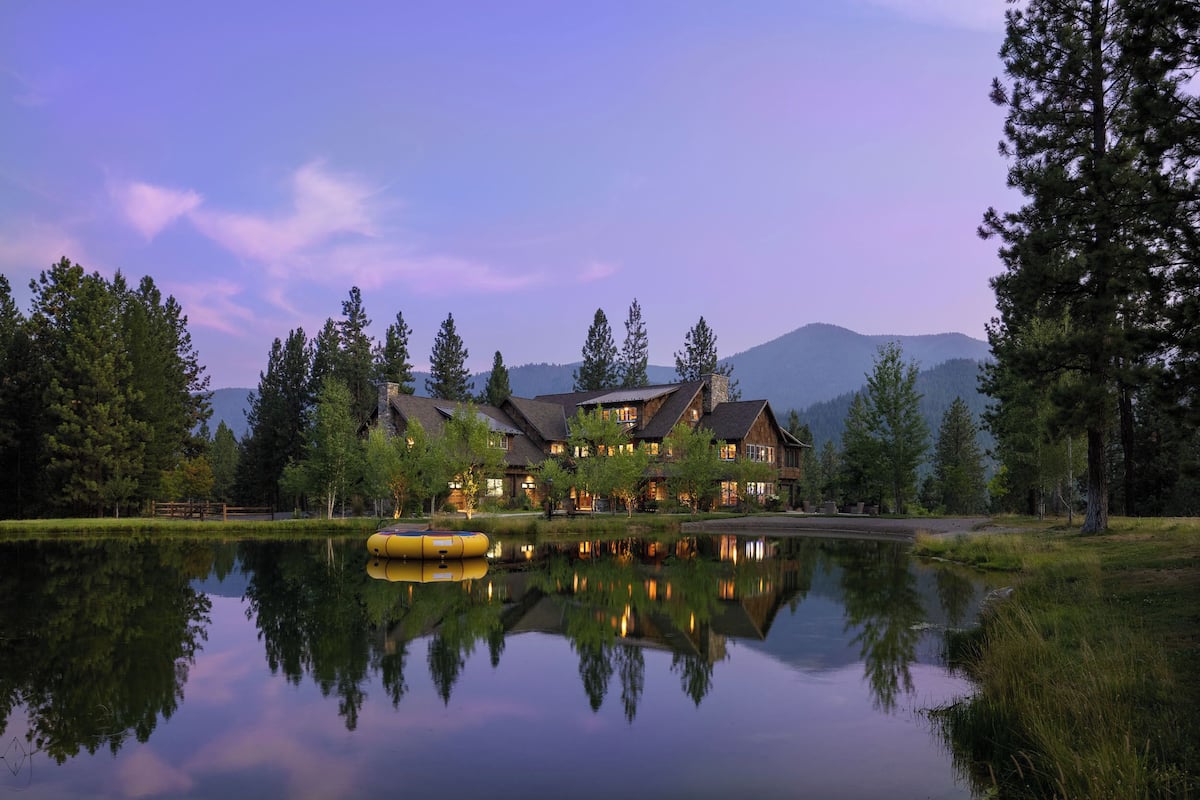 A large lodge with multiple buildings, a reflective lake, and surrounding trees.