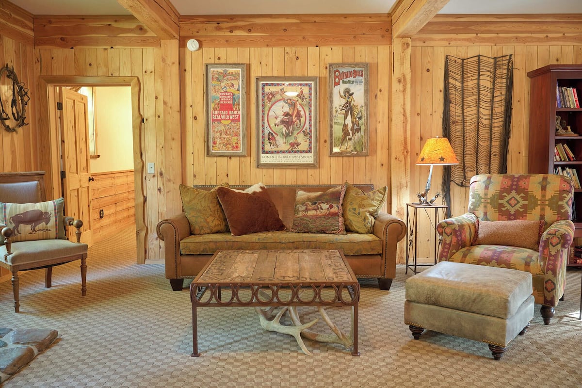 A cozy room with wooden walls, a sofa, a patterned armchair, a coffee table, and a side table with a lamp.