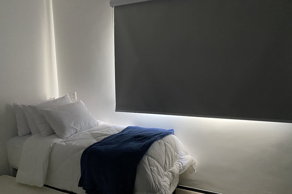 Une chambre à coucher avec un lit, du linge de lit blanc, une couverture bleue et une fenêtre avec un store.
