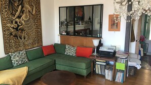 Een woonkamer met een groene modulaire bank, een houten salontafel, een kroonluchter, een boekenkast vol boeken, en een decoratief kunstwerk aan de muur.