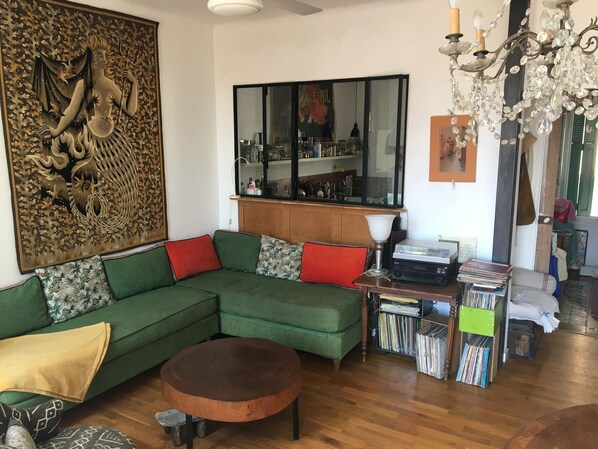 Un salon avec un canapé d’angle vert, une table basse en bois, un lustre, une bibliothèque remplie de livres et un élément décoratif mural.