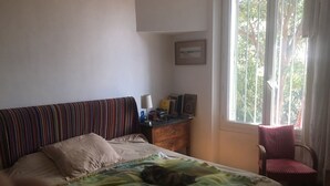 Een slaapkamer met een bed, een gestreept hoofdeinde, een raam met gordijnen, een stoel en een klein tafeltje met een lamp en boeken.