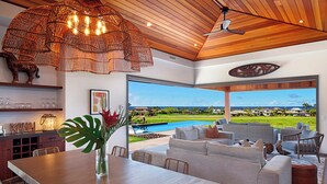 A modern living room with a wooden ceiling, a large dining table, and a view of a swimming pool and landscape. - Hale Kupanaha: Opulent New Villa + Pool/Spa/Views (Koloa)