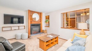 Una sala de estar moderna con chimenea, un televisor de pantalla plana montado en la pared, una mesa de centro de madera y un sofá con cojines.