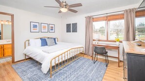 Un dormitorio con cama, escritorio, silla, ventilador de techo y ventana con cortinas.
