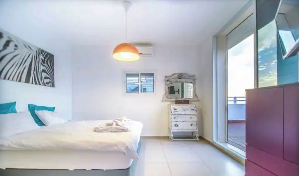 A bedroom with a bed, a ceiling fan, a window, a dresser, and a wall-mounted TV.