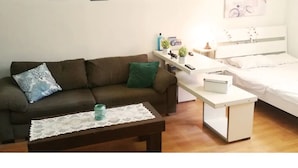 Une chambre meublée avec un canapé marron, une petite table recouverte d’une nappe en dentelle, une table de chevet blanche avec une lampe, et un lit avec une literie blanche.