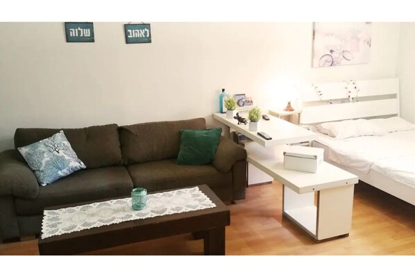 Een gemeubileerde kamer met een bruine bank, een klein tafeltje met een kantkleed, een witte nachtkastje met een lamp en een bed met witte beddengoed.