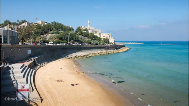 Une plage offrant une vue dégagée sur la mer, un rivage sablonneux et un bâtiment surmonté d’une tour, situé sur une colline en arrière-plan.