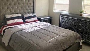A bedroom with a tufted headboard, a bed with a grey and white striped bedspread, a ceiling fan, and two windows with blinds. - Clearwater Home Away from Home (Clearwater)