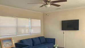 A living room with a dark blue sofa, a glass coffee table, a flat-screen TV mounted on the wall, and a ceiling fan. - Clearwater Home Away from Home (Clearwater)