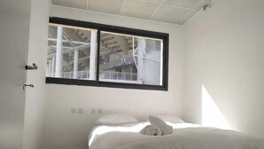 Quarto pequeno com uma cama de solteiro e janela com vista para uma obra.