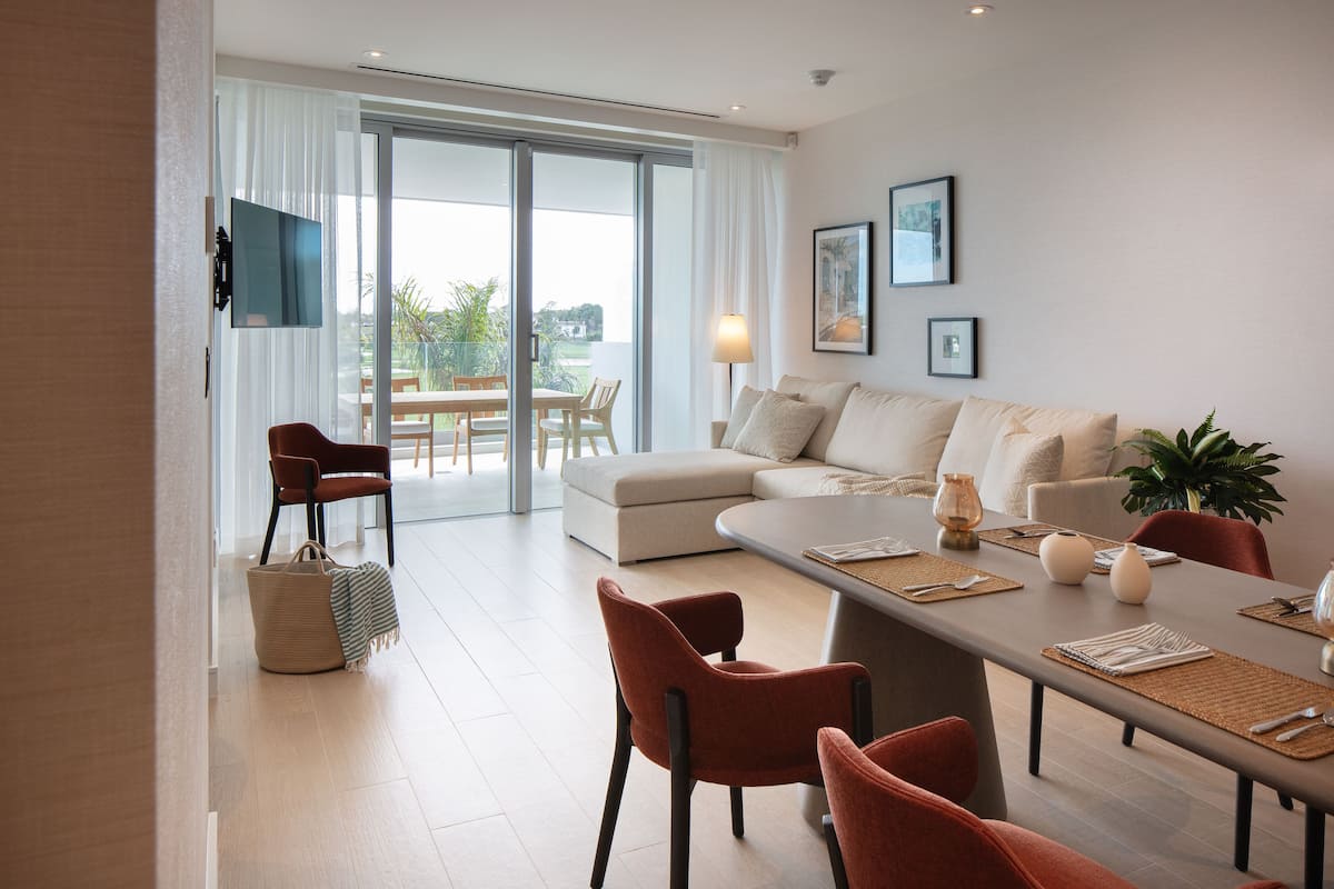 A modern living room with a sofa, dining table, and chairs. There is a TV mounted on the wall, a lamp, and a window with curtains.