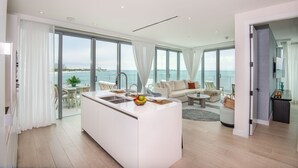 A modern living space with a kitchen island, dining area, and seating by a large window overlooking the sea. - Goldwynn Resort & Residences (Nassau)