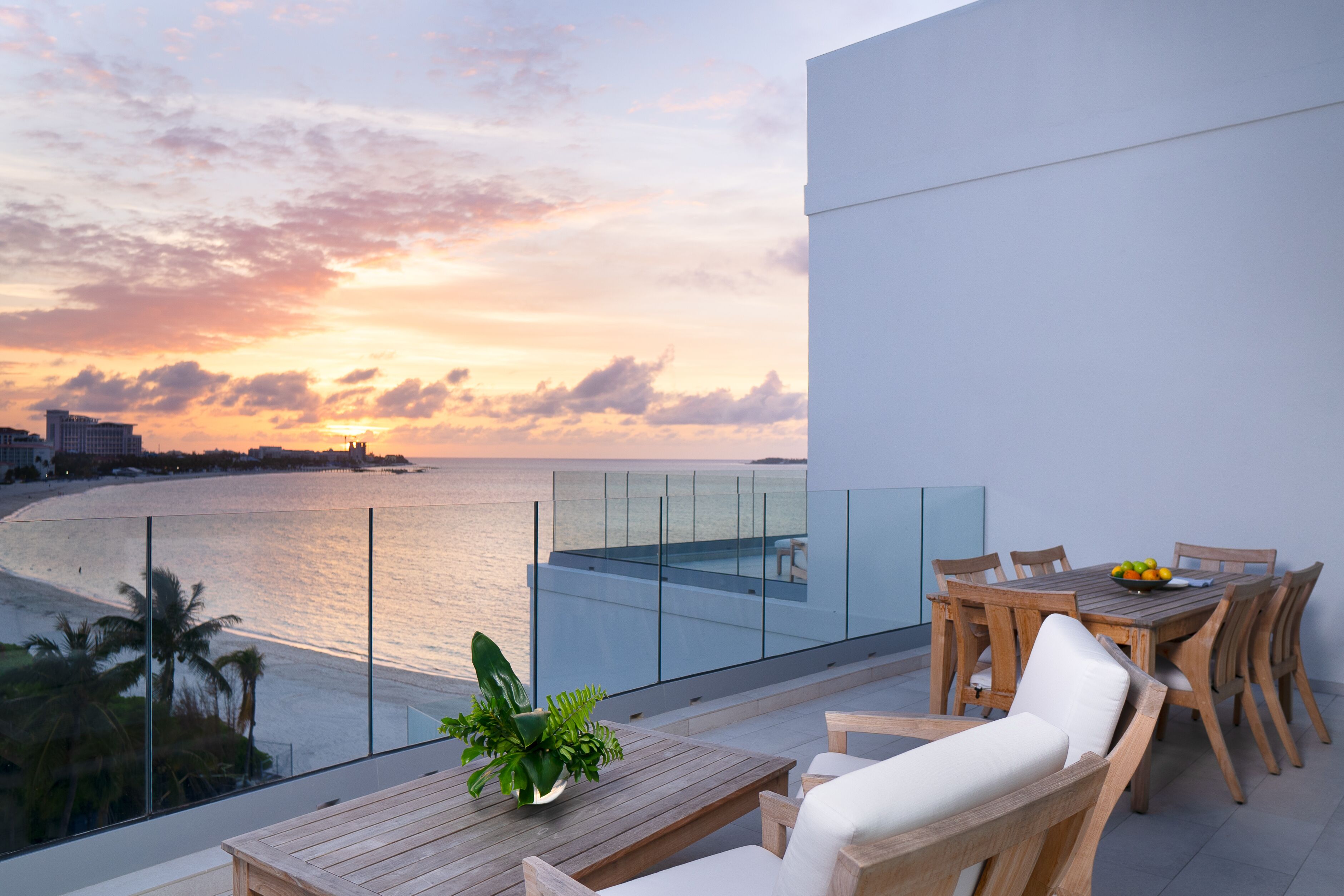 A balcony with a wooden table and chairs, overlooking a beach and ocean at sunset.