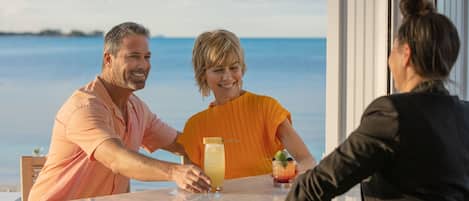 A bar area with a view of the sea, a glass table, and a person serving drinks.