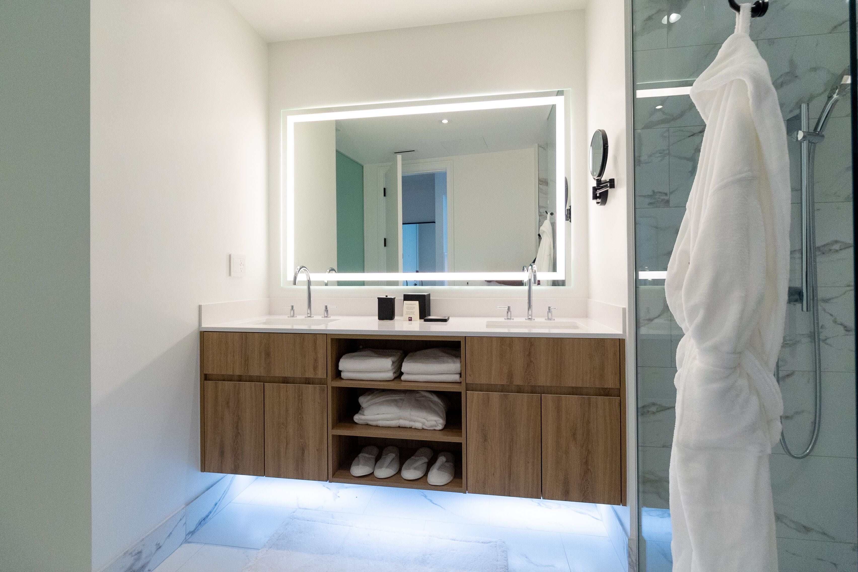 A modern bathroom with a large mirror, wooden vanity, shelves with towels and slippers, and a white robe hanging by the shower.